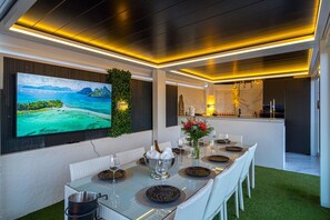 Een eetgedeelte met een lange tafel, stoelen, een aan de muur bevestigde televisie en een keuken op de achtergrond.