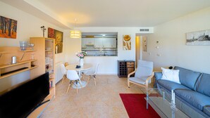 Un salon moderne avec un téléviseur à écran plat, un coin repas avec une table et des chaises blanches, un canapé bleu et une cuisine en arrière-plan.