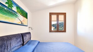 Une chambre à coucher avec un lit, un tableau représentant une scène de plage, une fenêtre donnant sur un beau paysage et une porte.