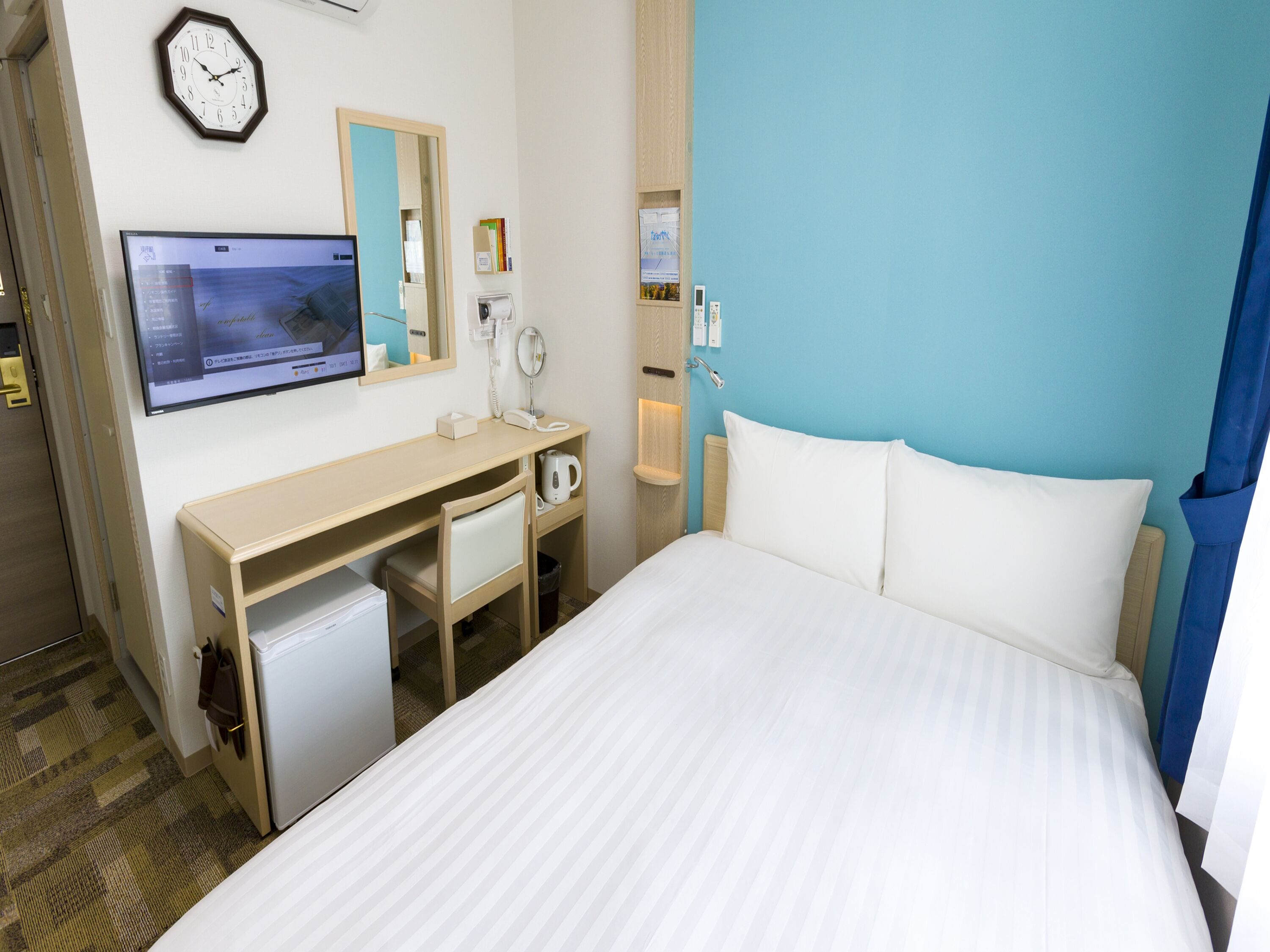 Une chambre d’hôtel équipée d’un lit, d’un bureau, d’une télévision, d’un miroir, d’une horloge et d’un téléphone fixé au mur.