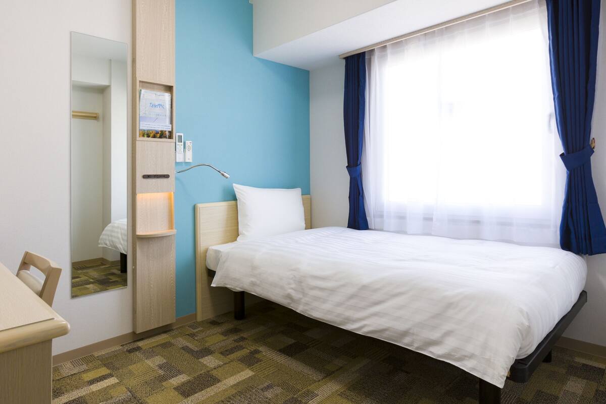 Ett hotellrum med en säng, ett sängbord, en spegel, en väggmonterad kontrollpanel och ett fönster med gardiner.