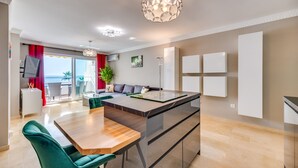 Eine moderne Küche mit einer zentralen Kochinsel, einem Essbereich mit Holztisch und grünen Stühlen sowie einer Wohnarea mit Sofa und Fernseher.