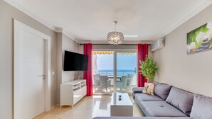 Ein modernes Wohnzimmer mit grauem Sofa, einem Flachbildfernseher, einem Couchtisch und einem Balkon mit roten Vorhängen.