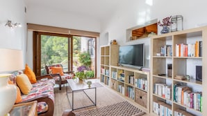 Een woonkamer met een bank, salontafel, boekenkast, televisie en uitzicht op groen.
