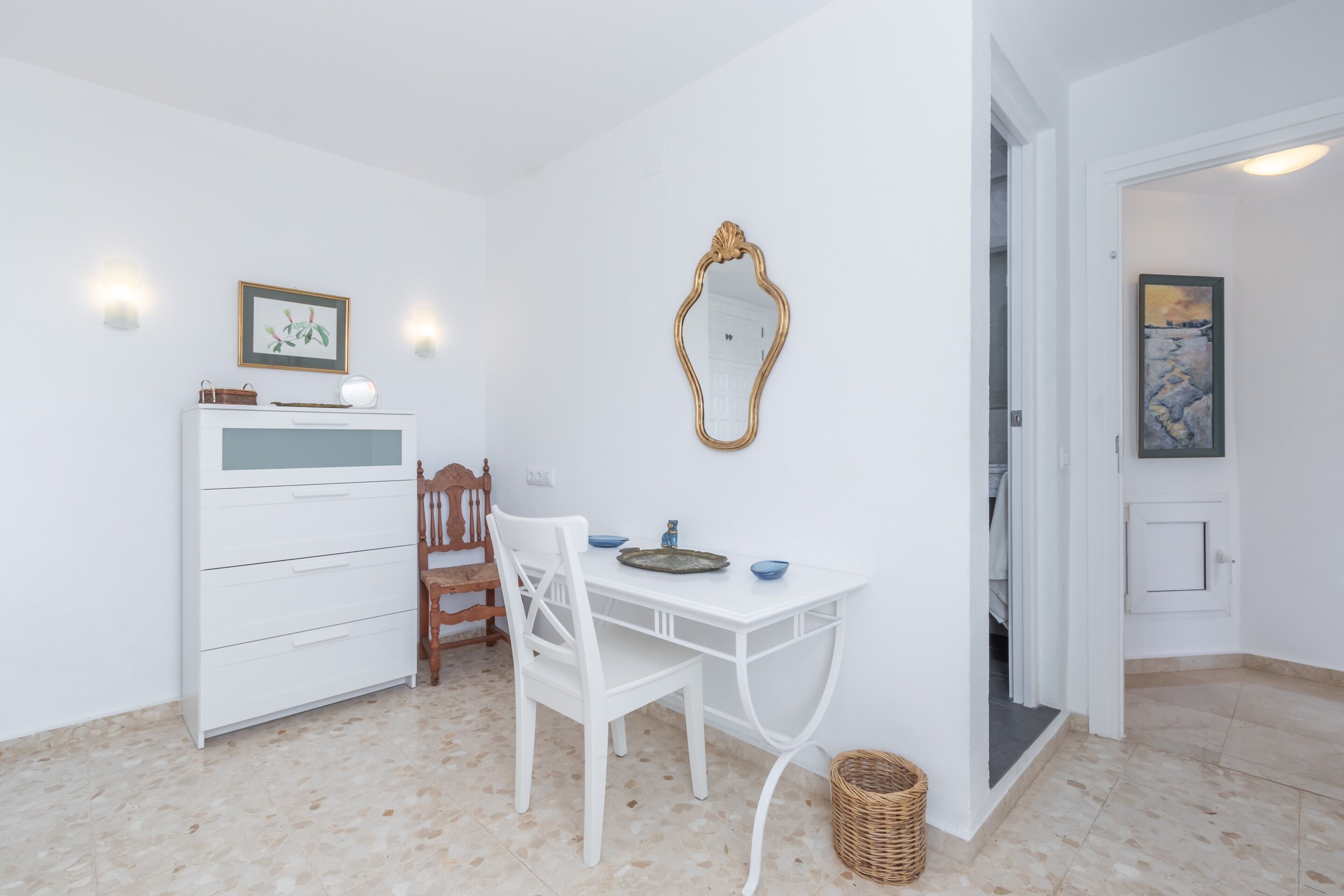 Une pièce blanche avec une chaise en bois, une table blanche, un miroir et un tableau encadré accroché au mur.