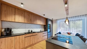 A modern kitchen with wooden cabinets, a central island, and a dining area with a large window. - UB31 Holiday Apartment – Exclusive Apartment with Private Wellness Oasis (Deutschnofen)