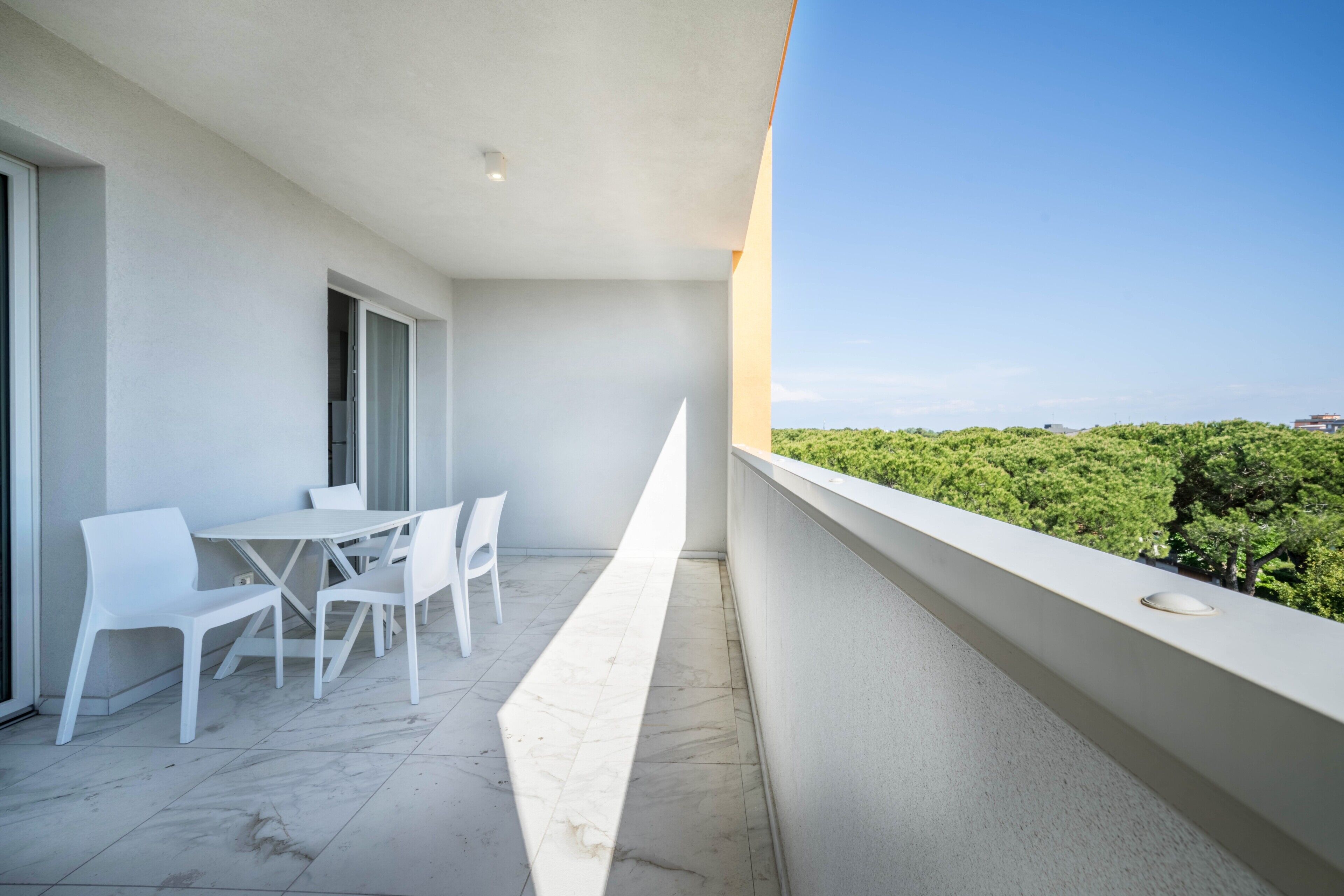 A balcony with a table and chairs, a view of greenery, and a clear sky.