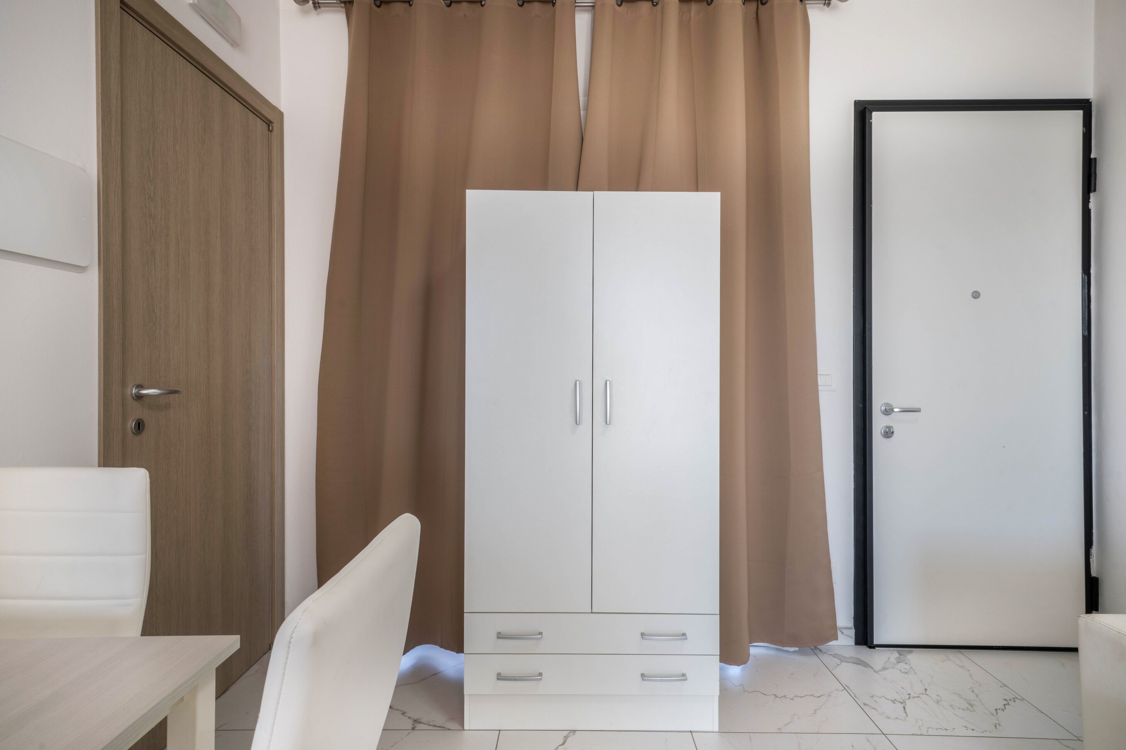 Una habitación con un armario blanco, una puerta de madera, una silla blanca y una puerta cerrada con manija.
