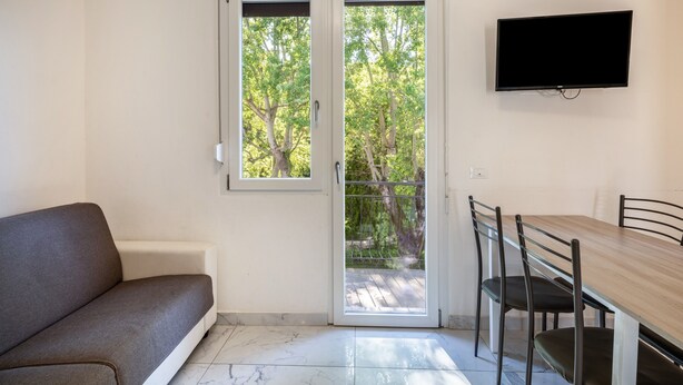 A room with a sofa, a dining table with chairs, a TV mounted on the wall, and a sliding glass door leading to an outdoor area with trees. - Holiday Apartment "Residenza Maja 6" with Wi-Fi (San Michele al Tagliamento)