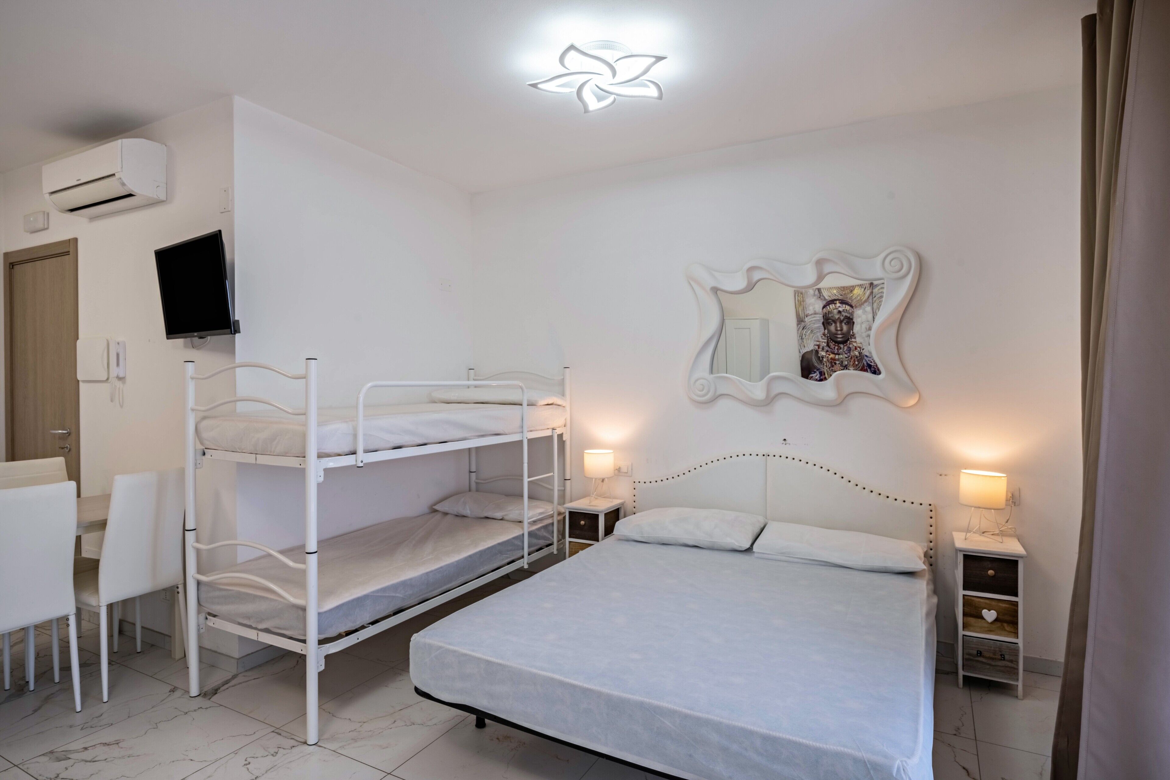 Quarto com beliches, escrivaninha, cadeira, mesa de cabeceira com abajur, TV na parede e espelho decorativo.