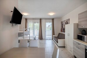 A modern hotel room with a flat-screen TV, a dining table, a sofa, a kitchen area, and a balcony with curtains.