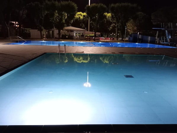 A swimming pool at night with a ladder leading into it, surrounded by trees and a building in the background. - Holiday Home - Villetta Cormor X with 3 Pools and Park (San Michele al Tagliamento)
