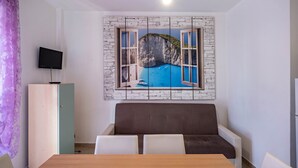 Une pièce comprenant une table à manger en bois, un canapé marron, un téléviseur fixé au mur et un tableau représentant un paysage côtier.