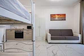 Une petite pièce comprenant un lit superposé, un coin cuisine, un canapé et un tableau représentant un coucher de soleil.