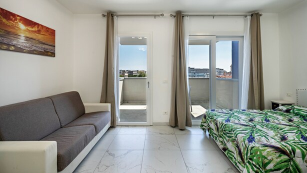 Habitación de hotel con sofá, cama con cubrecama floral, pintura de un atardecer y vista a través de puertas corredizas de vidrio.