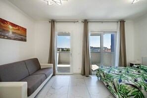 Une chambre d’hôtel avec un canapé, un lit recouvert d’une couvre-lit à motifs floraux, un tableau représentant un coucher de soleil et une vue à travers la porte-fenêtre coulissante.