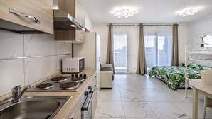 Una cocina moderna con microondas, estufa y pileta. Hay una cama con cubrecama estampado con hojas verdes y cabecera blanca.