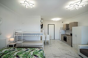 Une chambre de type dortoir, avec des lits superposés, un bureau, une chaise, une petite table avec une lampe, un coin cuisine et une porte.