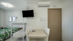 Una habitación con una cama individual y otra superior, una mesa con sillas, un televisor, aire acondicionado y una puerta de madera.