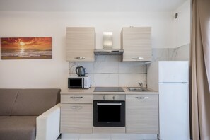 A compact kitchen with a microwave, stove, and refrigerator, a beige sofa, and a sunset painting on the wall.