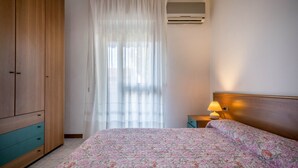 Un dormitorio con cama, un armario de madera, una mesita de noche con lámpara y una ventana con cortinas ligeras.
