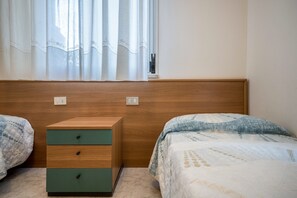 A bedroom with two beds, a wooden nightstand, and a window with curtains. - Appartamento della Bilancia 4 (Bibione)