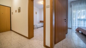 Una habitación con puerta de madera, cama con colcha estampada, suelo de baldosas y ventana con cortinas traslúcidas.
