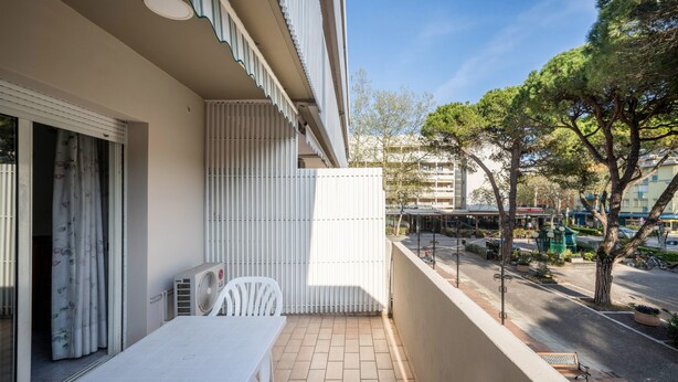 Un balcón con mesa y sillas, una ventana con persianas y vistas a una calle con árboles y edificios.