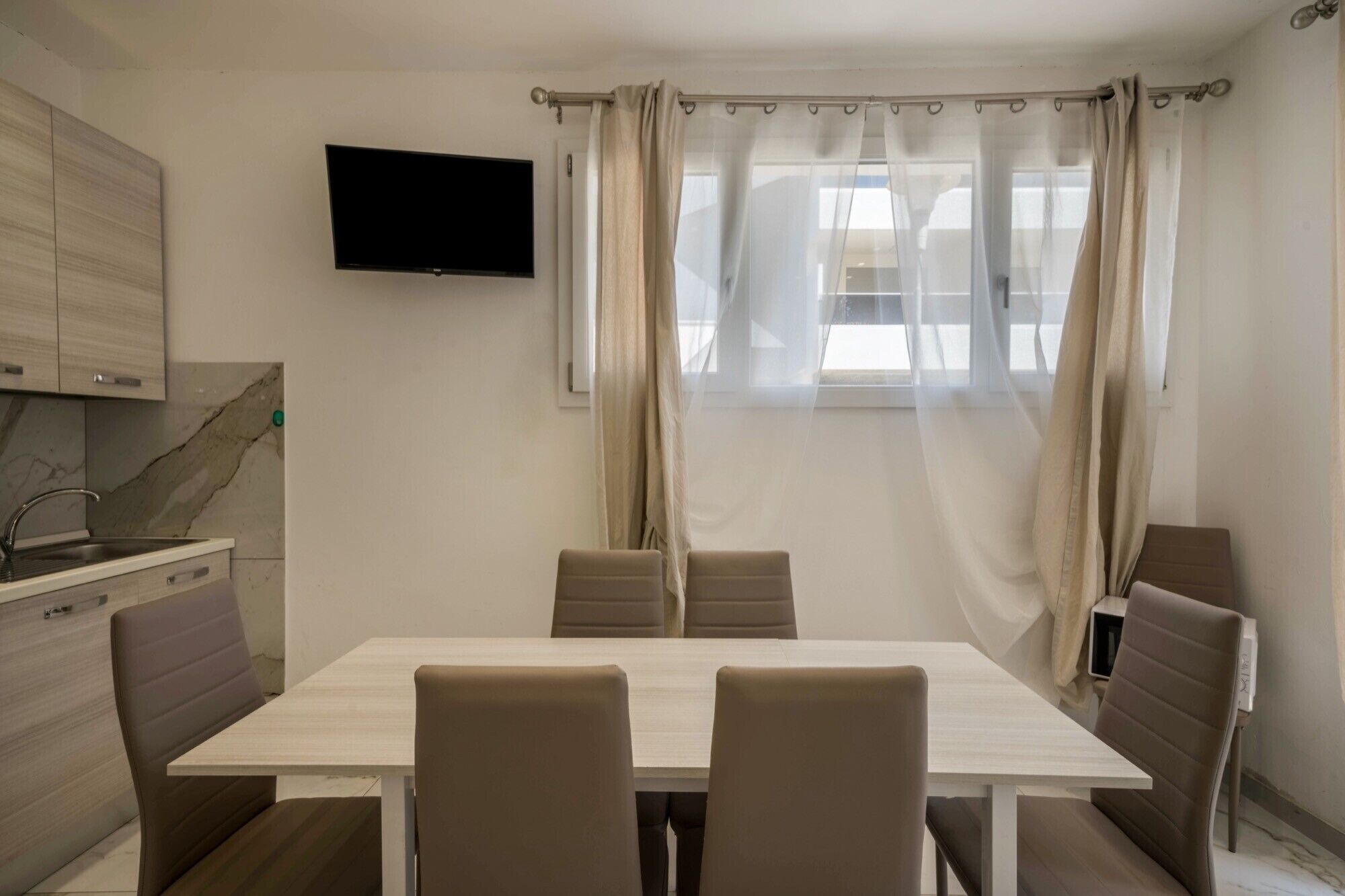 A modern kitchen with a flat-screen TV, a dining table with chairs, and a window with curtains.
