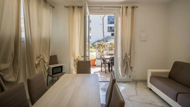 A room with a dining table, chairs, a sofa, a TV, and a view of a building and greenery outside. - Appartamento Andromeda 13 (Bibione)