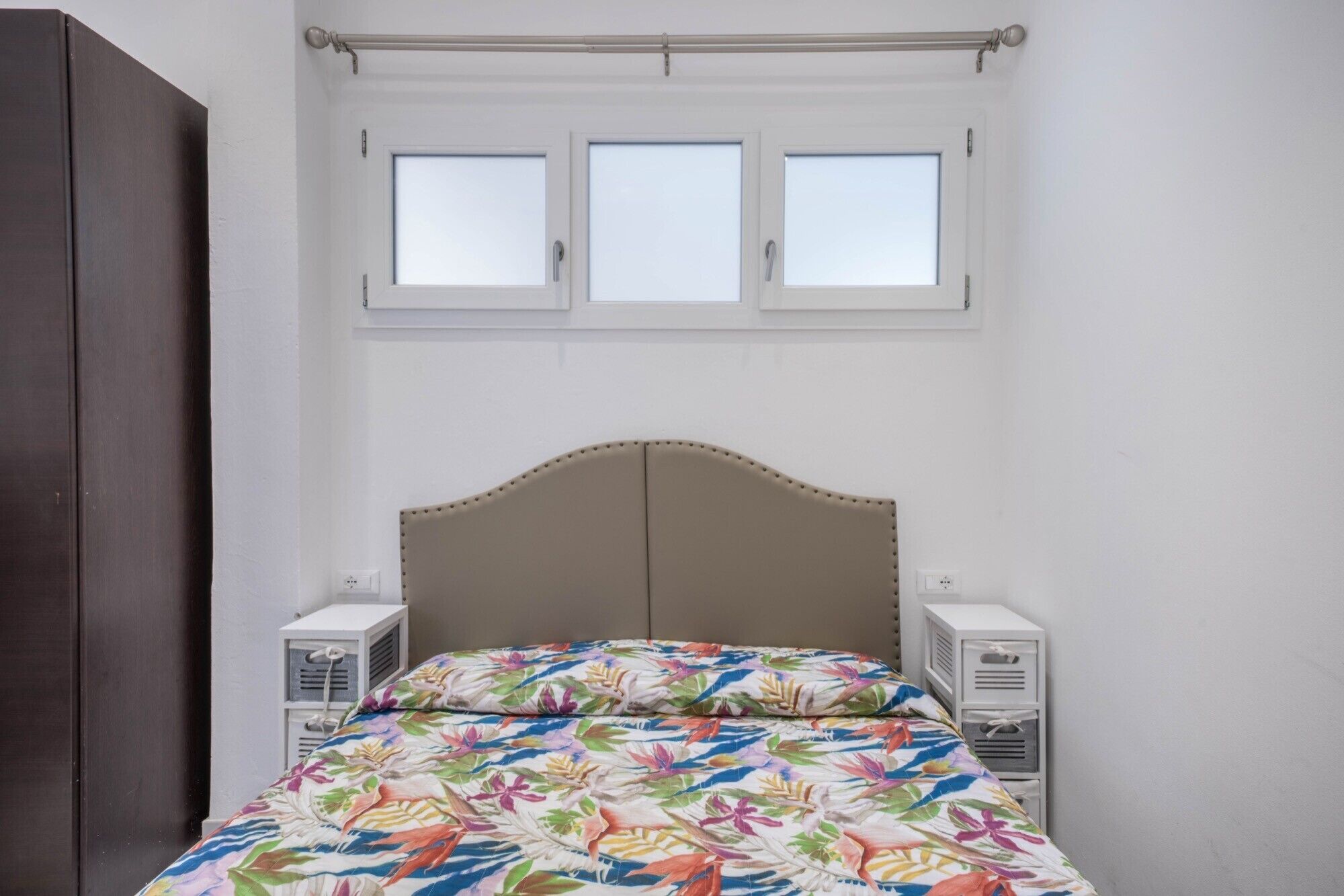 Een bed met een bloemenprint sprei, een houten garderobe en een raam met drie ruiten.