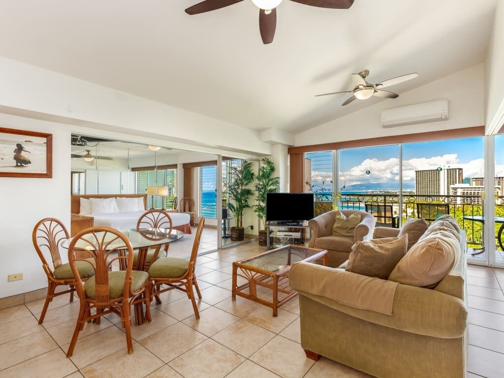 A spacious living room with a large window offering a city view, a dining area with wicker chairs, a sofa, and a flat-screen TV.