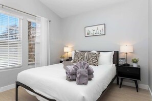 A neatly made bed with white bedding and two pillows, a bedside table with a lamp, and a window with blinds. - Modern 3 BR Home w/ Hot Tub, Pool, Golf! (North Las Vegas)