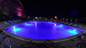 A nighttime swimming pool with blue lighting, surrounded by lounge chairs and a large rock formation.