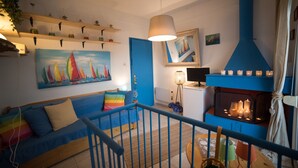 Ett vardagsrum med en blå soffa, en målning som föreställer segelbåtar, en öppen spis med ljus och en hängande lampa.