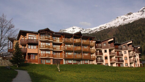 Un hotel de varios pisos con balcones, un camino y montañas al fondo.