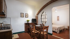 Eine kompakte Küche mit Gasherd, einem Esstisch mit Stühlen, einem Fernseher und einem kleinen TV-Schrank. Im angrenzenden Zimmer befindet sich ein Bett mit einer blumigen Tagesdecke.