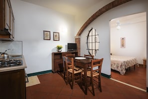 A compact kitchen with a gas stove, a dining table with chairs, a TV, and a small TV stand. There is a bed with a floral bedspread in the adjacent room.