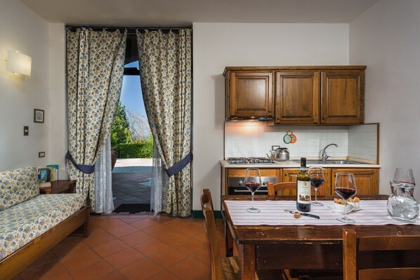 Une cuisine chaleureuse avec des meubles en bois, une table à manger dressée pour deux personnes et une vue sur l’extérieur à travers une fenêtre ornée de rideaux à motifs floraux.