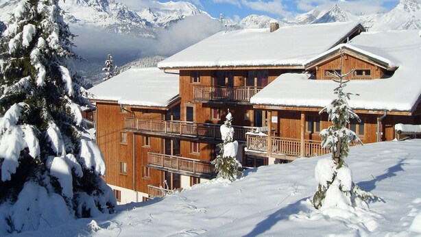 A wooden chalet with multiple balconies, surrounded by snow-covered mountains and trees. - Spacious 3 Rooms + Mountain Alcove, Sleeps 6/8, Close to Ski Lift and Shops (Valfréjus)