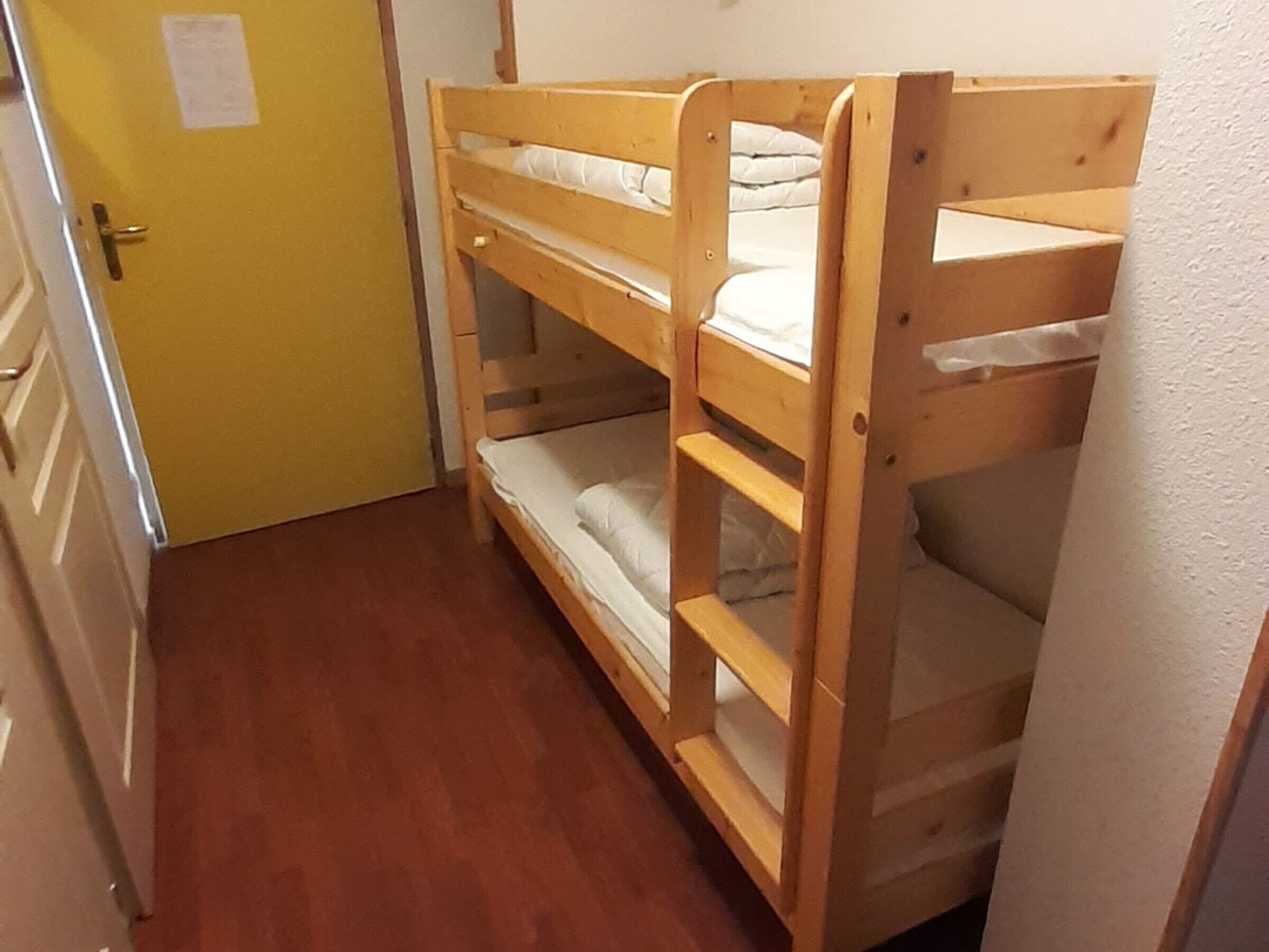 Una cama individual en un pasillo estrecho con suelo de madera y una puerta amarilla.
