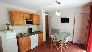 A compact kitchen with wooden cabinets, a microwave, a coffee maker, and a dishwasher. There is a glass dining table with chairs and a flat-screen TV mounted on the wall. - Spacious 3 Rooms + Mountain Alcove, Sleeps 6/8, Close to Ski Lift and Shops (Valfréjus)