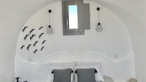 Un dormitori modern amb un sostre corbat, un llit gran amb roba de llit gris i blanca, una taula de nit amb una làmpada i una bandeja amb begudes i flors.