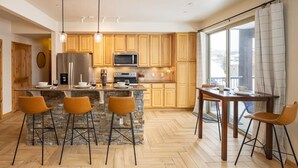 Eine moderne Küche mit Holzausstattung, einer zentralen Kochinsel inklusive Barhocker, einem Esstisch mit Stühlen und einem großen Fenster mit Vorhängen.