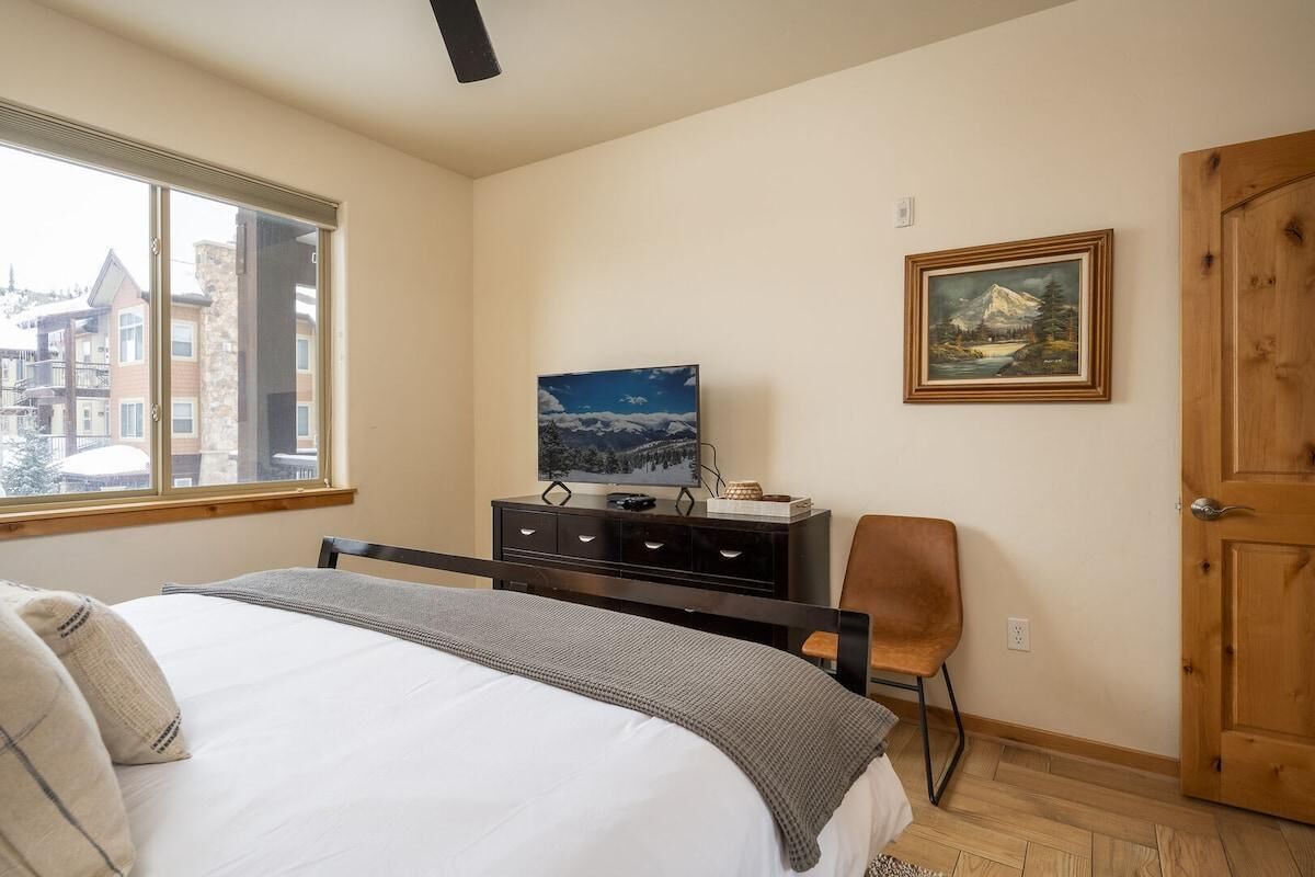 Une chambre à coucher avec un lit, un téléviseur posé sur une commode, une chaise, une porte et une fenêtre.