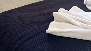 Una cama bien tendida con una colcha azul oscuro y toallas blancas, un cabecero gris y una mesita de noche con una lámpara.
