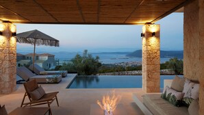 A luxurious outdoor seating area with a fire feature, overlooking a scenic view of the sea and hills.