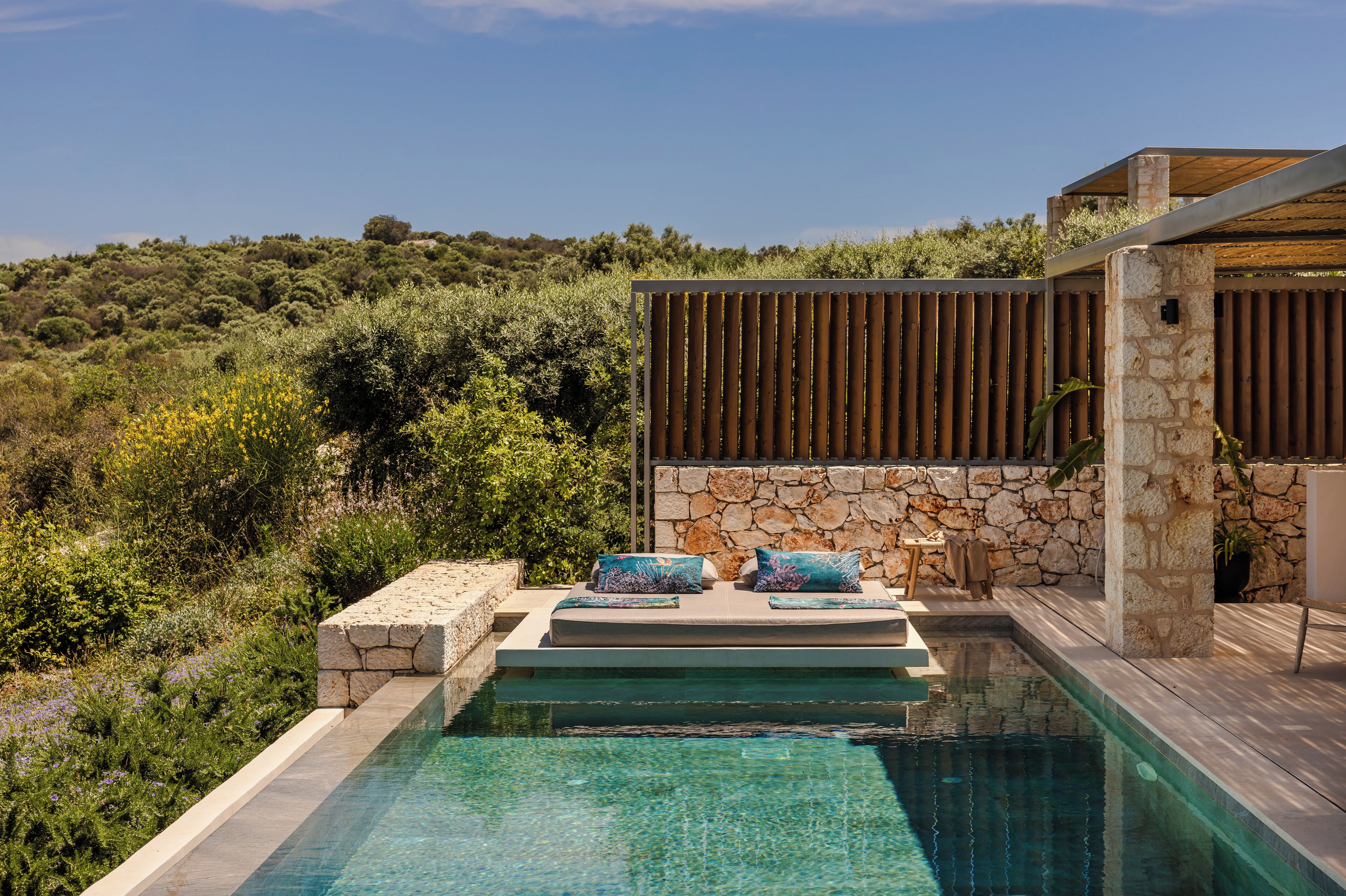 A pool with a stone wall and wooden slats, surrounded by lush vegetation and a view of a hillside.