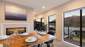 A dining area with a wooden table set for four, a fireplace, and large windows offering a view of a lake and mountains. - St Matthews Views - Cecil (Queenstown)