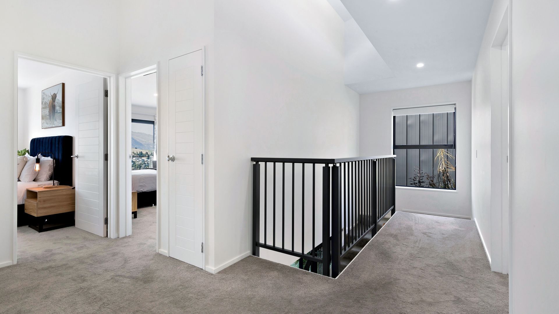 A modern hallway with a staircase, carpeted floors, and a view of a bedroom with a bed and a desk.